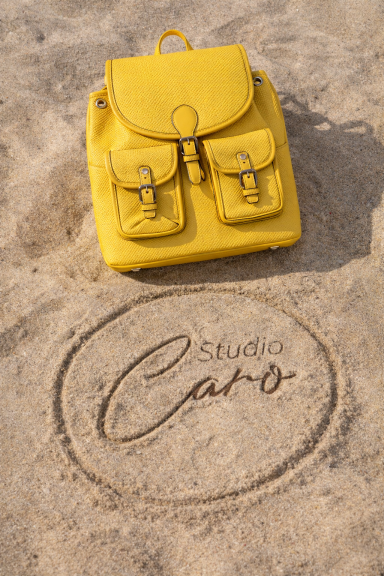 Sac à dos jaune posé sur du sable, avec "Studio Caro" écrit dans le sable.